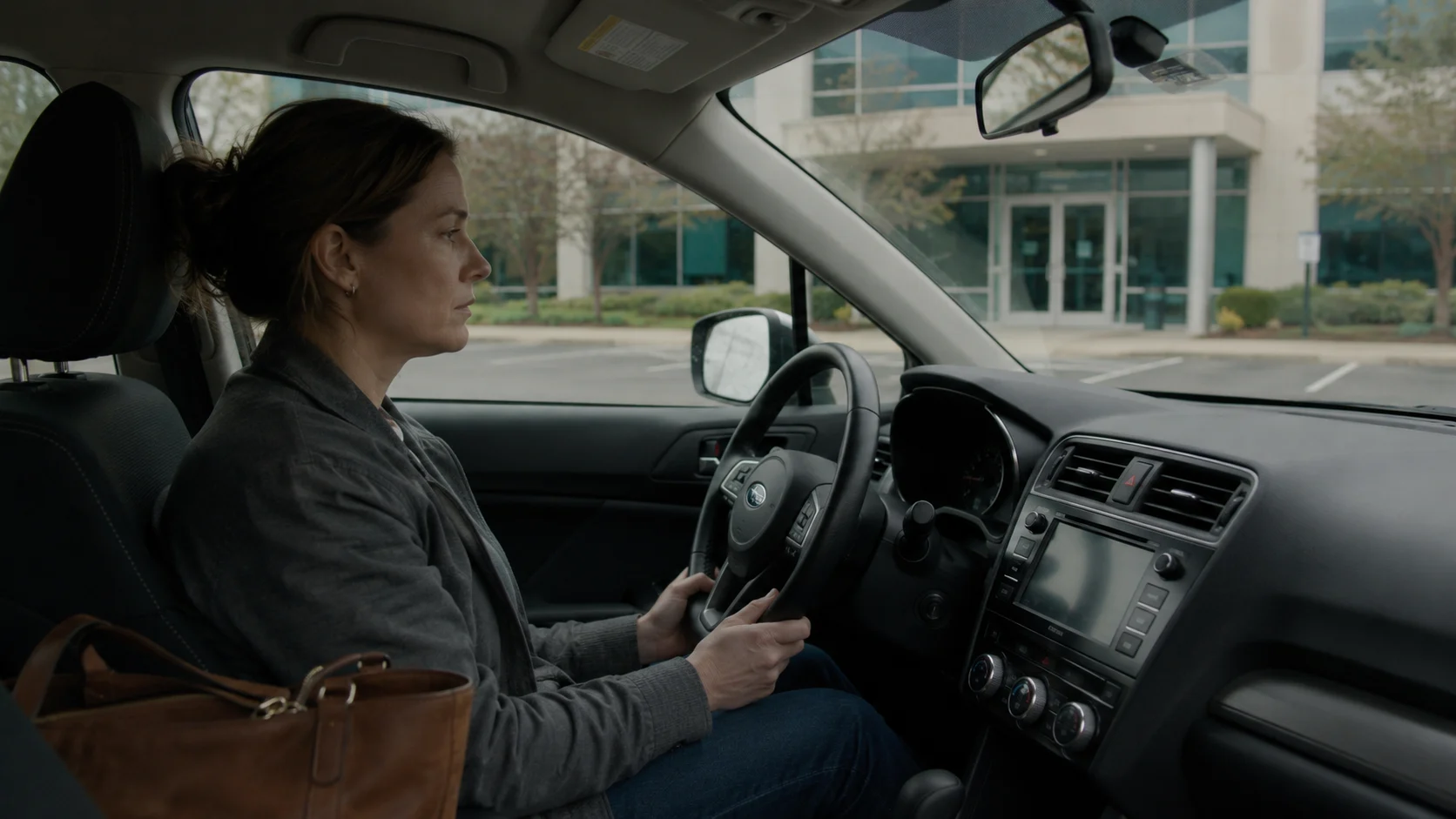 woman seated in parked car, hands on steering wheel, office building ahead
