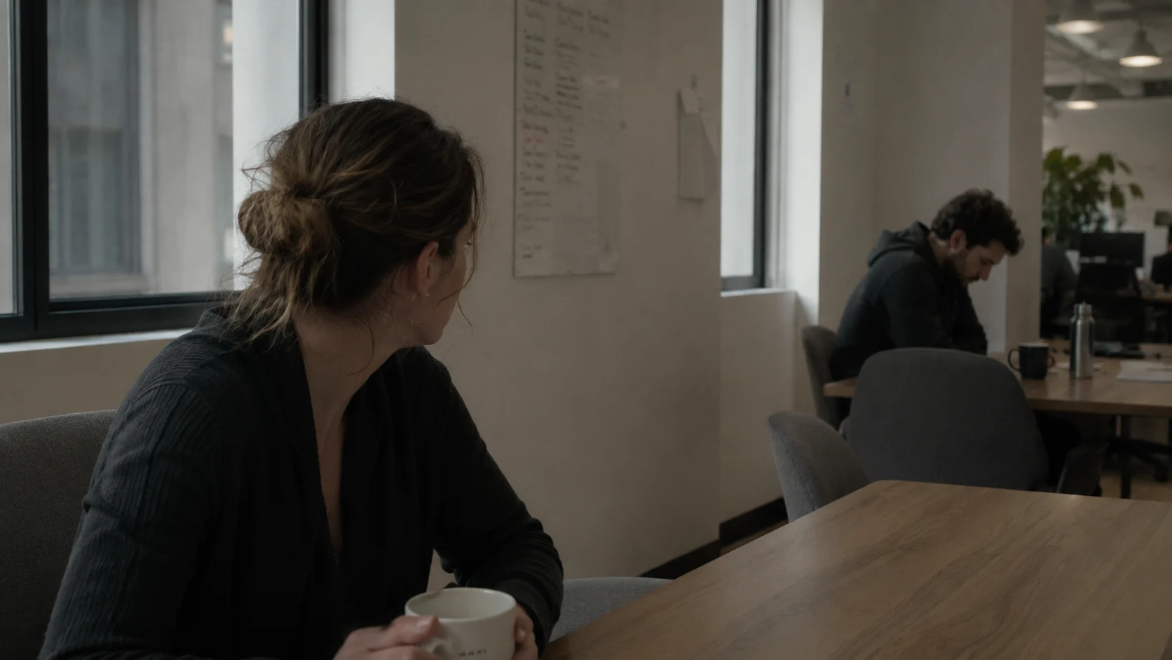 A person in an office pauses mid-turn toward a coworker sitting quietly in the background with a lowered head.