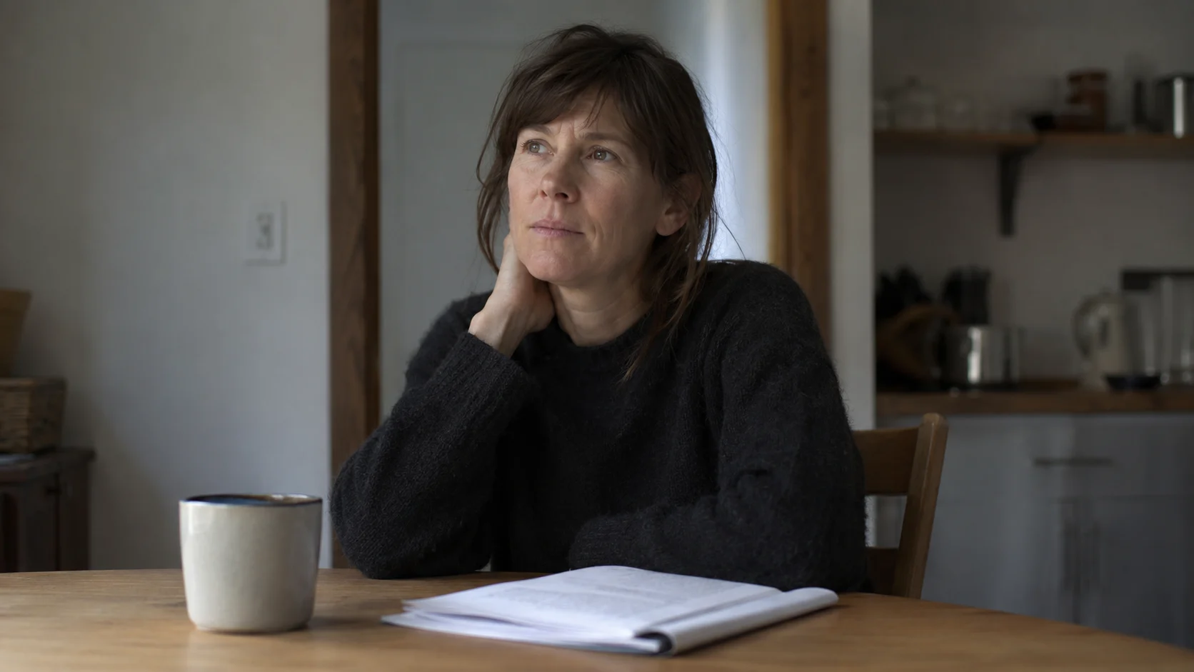 A person sitting alone at a kitchen table in flat morning light, mug untouched, looking inward with a quiet, arrested expression, a moment of private realization.